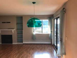 View from my dining area looking towards my lounge with a low-hanging semi-transparent green light shade, light brown walls, and dark brown laminate flooring. A double window in the background.