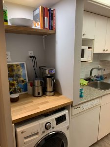 Photo of laundry area after reonvation with washer/dryer in place, countertop above, and a shelf. Plus fresh paint. In the space is a coffee machine, milk frother, grinder, a photo of sunflowers and books on a shelf.